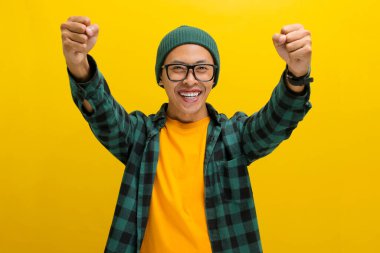 Asyalı heyecanlı genç bir adam, bereli bir şapka ve günlük bir gömlek giyerek, sarı bir arka planda dururken sevinç ve zaferi bir yumruk pompasıyla ya da EVET hareketiyle kutluyor.