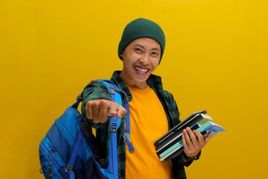 Şapka ve gündelik gömlek giyip sırt çantası taşıyan neşeli Asyalı genç bir öğrenci, elinde bir kitap tutuyor ve kameraya şevkle işaret ediyor, seçim, davetiye ya da işe alım işareti veriyor.