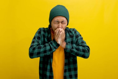 Şapka ve günlük gömlek giyen genç Asyalı bir adam, muhtemelen sarı arka planda izole edilmiş soğuk algınlığı veya alerjiden dolayı burnunu mendile sümkürürken görülüyor. Sağlık hizmetleri ve tıbbi kavramlar