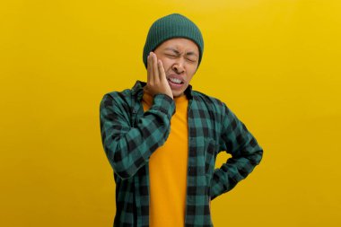 Şapkalı ve günlük gömlekli genç Asyalı bir adam diş ağrısı çekiyor, dişinde rahatsızlık ya da ağrı hissediyor, sarı arka planda izole edilmiş. Sağlık hizmetleri ve tıbbi kavramlar