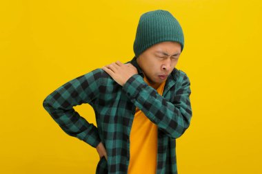 Şapka ve günlük kıyafet giymiş Asyalı bir adam omzuna masaj yaparken görülüyor. Aşırı çalışma nedeniyle sırt ağrısı ve omuz ağrısı şikayeti var. Sarı arka planda izole edilmiş..