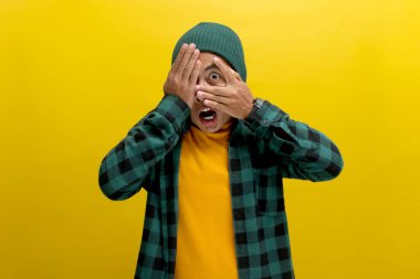 Şapka ve gündelik gömlek giymiş, şaşırmış genç bir Asyalı adam, elleriyle yüzünü ve gözlerini kapatıyor, şok olmuş ve utanmış bir ifadeyle parmaklarının arasından bakıyor.