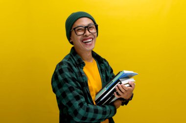 Genç bir Asyalı öğrenci, gözlüklü, bereli, sıradan elbiseli, sarı arka planda duruyor, göğsüne kitap dayamış..