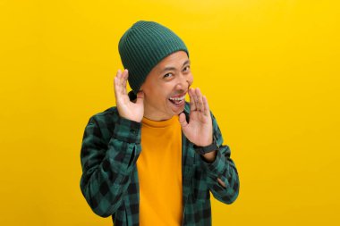 Young Asian man, dressed in a beanie hat and casual shirt, laughs while listening intently, hand cupped over his ear, possibly catching wind of rumor or gossip while standing against yellow background clipart