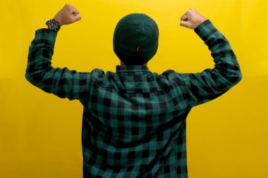 Kendine güveni tam Asyalı bir adam, bereli bir şapka ve sıradan bir gömlek giymiş, kolunu kaldırıp kaslarını göstererek güçlü bir jest yapıyor, başarılarından gurur duyuyor, sarı arka planda duruyor.