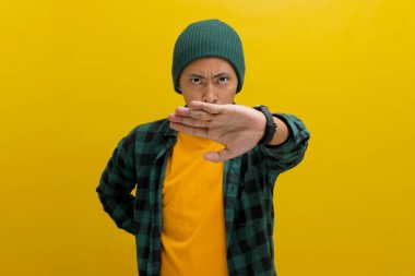 Hoşnut olmayan genç Asyalı bir adam, bereli bir şapka ve gündelik bir gömlek giyerek, kollarını kameraya doğru uzatarak, sarı bir arkaplana karşı durarak bir ret ya da dur hareketi sergiliyor.