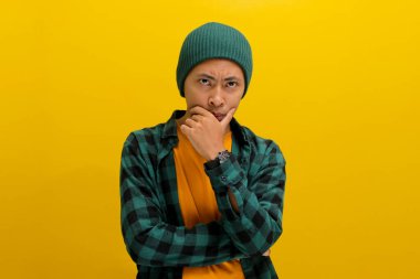 Şapka ve gündelik gömlek giyen düşünceli genç Asyalı adam, elini çenesine koyar, kafası karışmış bir ifadeyle kameraya bakar, derin bir şekilde düşüncelere dalmış ve geleceği düşünür.