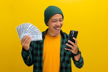 Asyalı heyecanlı bir adam bereli ve günlük kıyafetlerle telefonuna bakarken şaşırmış bir ifadeyle sarı arka planda izole edilmiş para tutuyor..