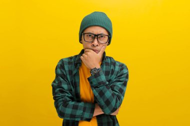 Şapka ve günlük gömlek giyen düşünceli genç Asyalı adam, elini çenesinin üzerine koyup, sarı arka planda dururken gelecek hakkında düşünürken endişeli görünüyor..