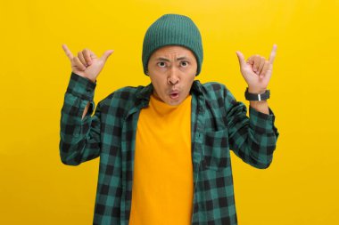 Şapka takan ve günlük kıyafetler giyen genç Asyalı bir adam, kameraya bir kaya jesti ve gülümsüyor, en sevdiği grup konserine katılmaya hazır olduğunu gösteriyor, sarı arka plan