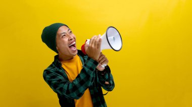 Bereli ve rahat gömlekli heyecanlı Asyalı adam, haberleri, özel bir teklifi, indirimi veya satışı paylaşmak için hoparlörü kullanarak megafonla bir duyuru yapıyor. Sarı arka planda izole edilmiş..