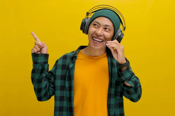 stock image Excited Asian man in a beanie and casual clothes, wearing headphones and enjoying music or a podcast, points excitedly towards empty copy space with a happy expression. Isolated on a yellow background