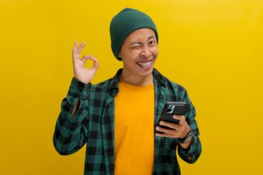 Asyalı heyecanlı bir genç adam, bereli bir şapka ve günlük bir gömlek giyer, kameraya doğru gözünü kırpar ve kameraya doğru iyi bir hareket yapar, akıllı telefonunu tutar, onayını iletir ve memnuniyetini sunar.