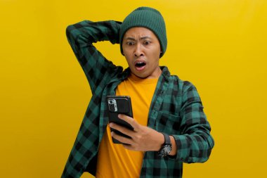 Şaşkın Asyalı bir adam, bereli bir şapka ve gündelik bir gömlek giyer, telefonunda sarı bir arka plana karşı durarak özel bir indirime şaşırır.