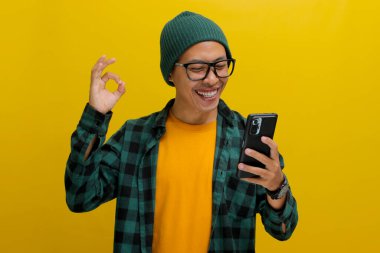 Şapka ve günlük kıyafetler giyen Asyalı genç bir adam, akıllı telefonunu tutarken, anlaşma, onay ve memnuniyet iletirken, olumlu bir değerlendirmeyi işaret ederken, iyi bir işaret hareketi yapıyor.
