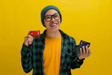 Asyalı heyecanlı genç bir adam, bereli şapka ve günlük gömlek giymiş, bir cüzdan ve kredi kartı tutuyor, kameraya mutlulukla gülümsüyor, kredi kartını kullanarak online ödeme yapmaya hevesli.