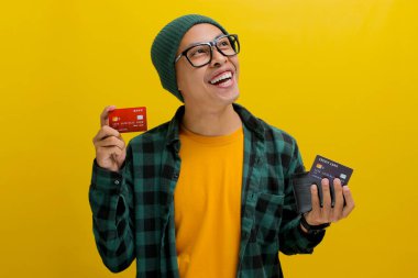 Asyalı heyecanlı genç bir adam, bereli şapka ve günlük gömlek giymiş, bir cüzdan ve kredi kartı tutuyor, kameraya mutlulukla gülümsüyor, kredi kartını kullanarak online ödeme yapmaya hevesli.