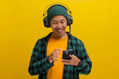 Asian man in a beanie and casual clothes browses a music app on his phone, selecting a song from a music app while wearing headphones. Isolated on a yellow background. clipart