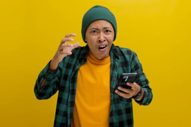 Şapka ve günlük gömlek giyen mutsuz Asyalı adam, akıllı telefonunu tutarken şaşırmış ve üzgün görünüyor, muhtemelen kötü haberlere tepki veriyor ya da online dolandırıcılara kurban gidiyor.