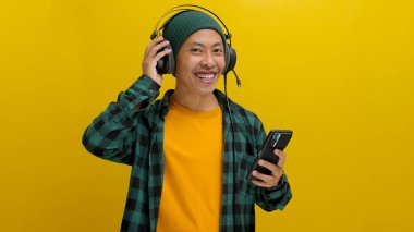 Asian man in a beanie and casual clothes listens to music on his headphones, phone held in hand. Isolated on a yellow background. clipart