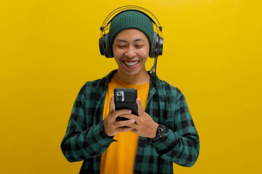 Asian man in a beanie and casual clothes browses a music app on his phone, selecting a song from a music app while wearing headphones. Isolated on a yellow background. clipart