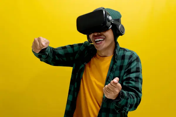 stock image Young Asian man wearing a VR headset races a virtual car, gripping an imaginary steering wheel. Isolated on a yellow background