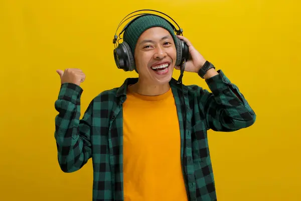 stock image Excited Asian man in a beanie and casual clothes, wearing headphones and enjoying music or a podcast, points excitedly towards empty copy space with a happy expression. Isolated on a yellow background