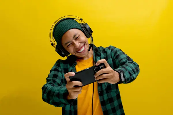 stock image Excited young Asian man, wearing headphones, a beanie hat and casual shirt, engages in playing an online mobile game on his phone while simultaneously live broadcasting the gameplay on the internet