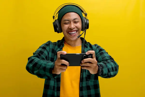 Stock image Excited young Asian man, wearing headphones, a beanie hat and casual shirt, engages in playing an online mobile game on his phone while simultaneously live broadcasting the gameplay on the internet