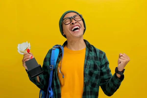 A clever Asian male student, dressed in a beanie hat and casual attire, raises his fist in a YES gesture, celebrating the win of a trophy, rejoicing in his success and accomplishments