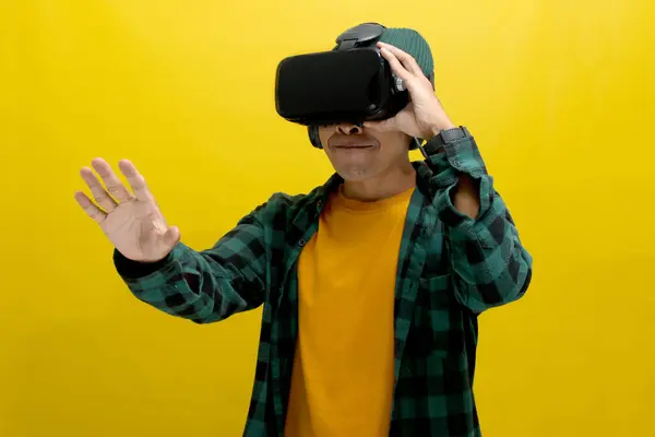 stock image Excited Asian man wearing a VR headset reaches out with his hand, seemingly interacting with a virtual object in a VR game. Isolated on a yellow background