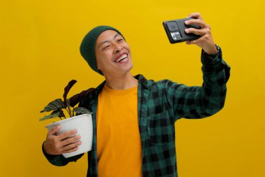 Cheerful Asian man in a beanie and casual clothes takes a selfie with his healthy Pin-stripe Calathea (Calathea ornata) houseplant in a white pot. Isolated on a yellow background. clipart