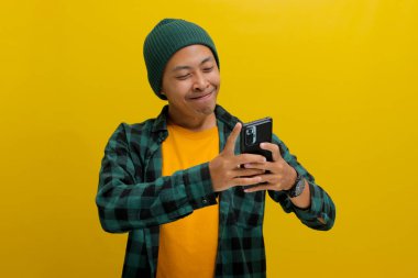Şapka ve günlük gömlek giyen mutlu Asyalı adam, akıllı telefonunu tutuyor, sarı bir arka planda dururken neşeli bir gülümseme sergiliyor.