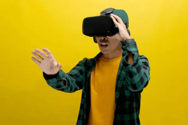 stock image Excited Asian man wearing a VR headset reaches out with his hand, seemingly interacting with a virtual object in a VR game. Isolated on a yellow background