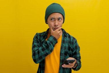 Şapka ve gündelik tişört giyen düşünceli Asyalı bir adam, elinde bir cep telefonu tutuyor, eli çenesinde ciddi ve derin düşünceler taşıyor, mesaj okurken şüphe ve endişe gösteriyor.