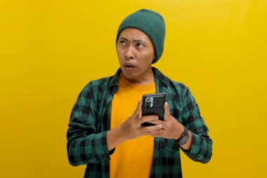 Şapka ve günlük gömlek giyen mutsuz Asyalı adam, akıllı telefonunu tutarken şaşırmış ve üzgün görünüyor, muhtemelen kötü haberlere tepki veriyor ya da online dolandırıcılara kurban gidiyor.