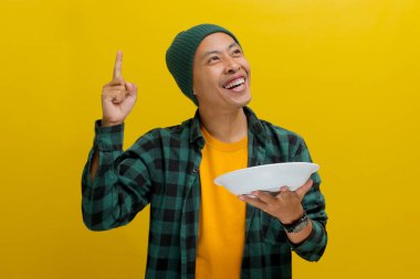Şapka ve günlük gömlek giyen genç Asyalı adam, boş beyaz bir tabak tutarken parmağını kaldırıyor, kameraya gülümsüyor ve sarı bir arkaplanda duruyor.
