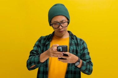 Şapka ve günlük gömlek giyen Asyalı bir adam, haberlere, mesajlara ya da telefonundaki e-posta ve sosyal medyaya bakarken, sarı arka planda izole edilmiş bir şekilde gözlüğünü ayarlıyor.