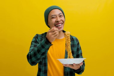 Asian man in a beanie and casual clothes enjoys slurping instant noodles using fork. Isolated on a yellow background. clipart