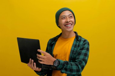Kendine güvenen ve neşeli Asyalı işadamı, bereli şapka ve günlük gömlek giymiş, elinde bir dizüstü bilgisayarla boş bir fotokopi odasına bakıyor, kendine güven, mutluluk ve parlak bir gülümseme, sarı bir arka plan.