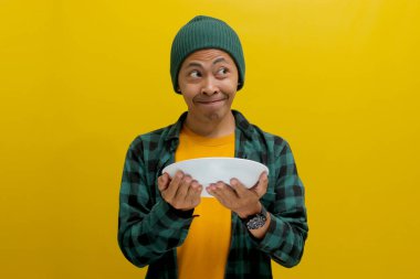 Aç genç Asyalı bir adam elinde bir tabak tutarken boş bir fotokopi alanına doğru bakıyor, sarı bir arka planda dururken, heyecanlı bir şekilde ne yiyeceğini düşünüyor.