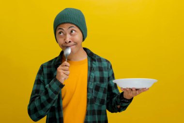 Şapkalı ve sıradan gömlekli düşünceli genç Asyalı bir adam bir kaşık ve boş beyaz bir tabak tutuyor, sarı arka planda dururken ne yiyeceğini düşünüyor.