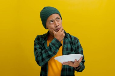 Şapka ve gündelik gömlek giyen düşünceli genç Asyalı adam elinde boş beyaz bir tabak tutarken ne yiyeceğini düşünüyor ve yemek pişirme menüsünü düşünüyor.