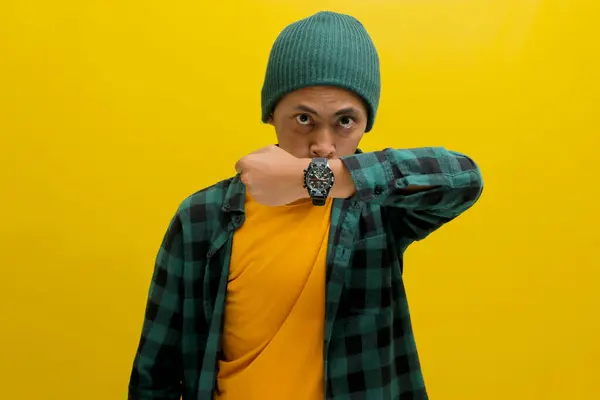 Impatient Asian man in a beanie and casual shirt shows his watch to the camera, reminding about a schedule, target, or deadline. Isolated on a yellow background