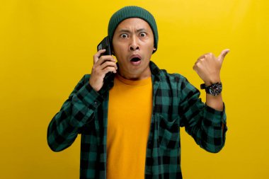 Şok olmuş Asyalı bir adam, bereli bir şapka ve günlük bir gömlek giyer, boş bir yere başparmağıyla işaret eder, cep telefonuyla konuşurken mesaj veya reklam için mükemmeldir.