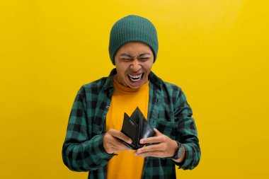 Şok olmuş genç Asyalı bir adam, bereli bir şapka ve sıradan bir gömlek giymiş, boş cüzdanına kızgın bir şekilde bakıyor, iflasa yol açabilecek mali zorluklarla karşı karşıya, sarı bir arka planda izole edilmiş.