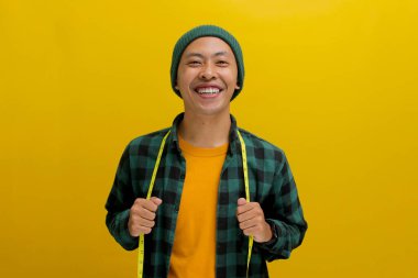 Cheerful Asian man in a beanie and casual shirt smiles at the camera, holding a yellow measuring tape around his neck. Isolated on a yellow background clipart