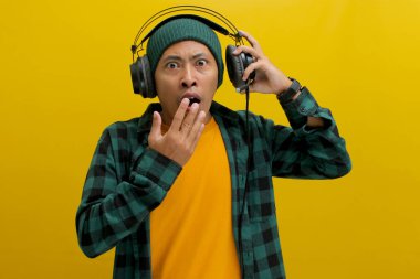 Asian man in a beanie and casual clothes, wearing headphones, looks shocked with his mouth agape after hearing something surprising on his music. Isolated on a yellow background. clipart