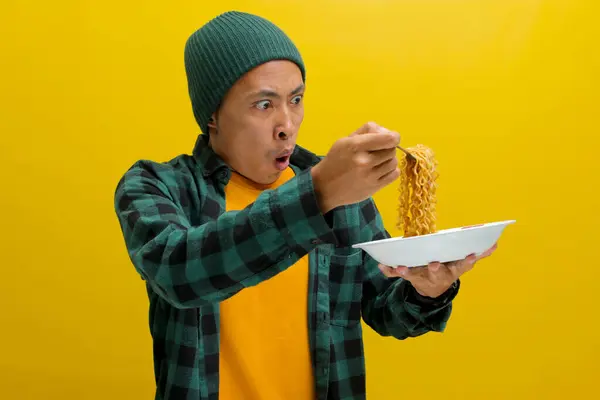 stock image Asian man in a beanie and casual clothes makes a surprised and delighted expression while slurping instant noodles with Fork. Isolated on a yellow background.