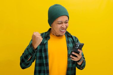 Öfkeli Asyalı bir adam, bereli bir şapka ve sıradan bir gömlek giyer, kötü haberlere görünür bir şekilde tepki verir, cep telefonuna bakarken, sarı arka planda durur.